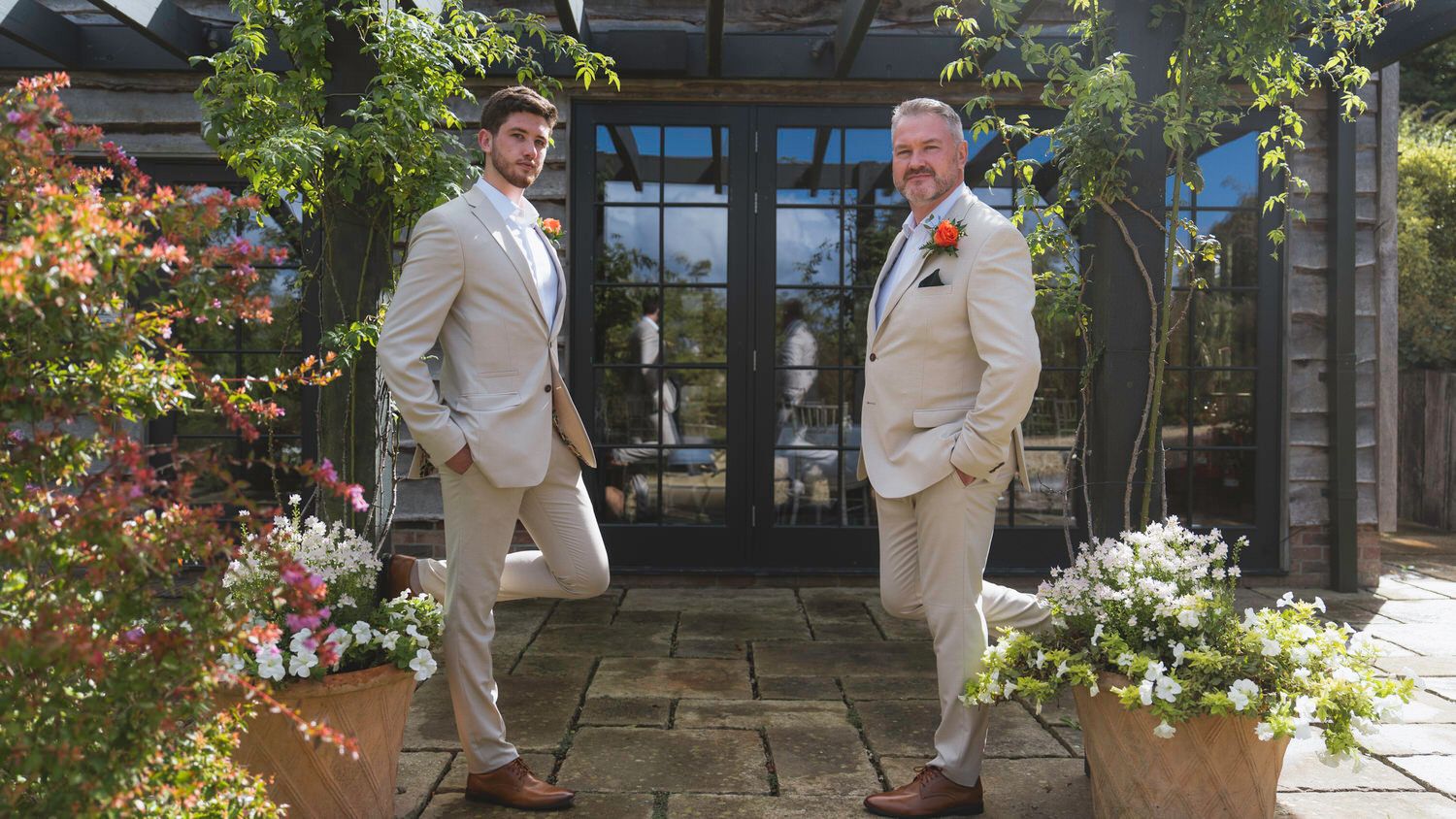 Two grooms in matching beige suits pose outdoors among colorful flowers at a wedding venue.