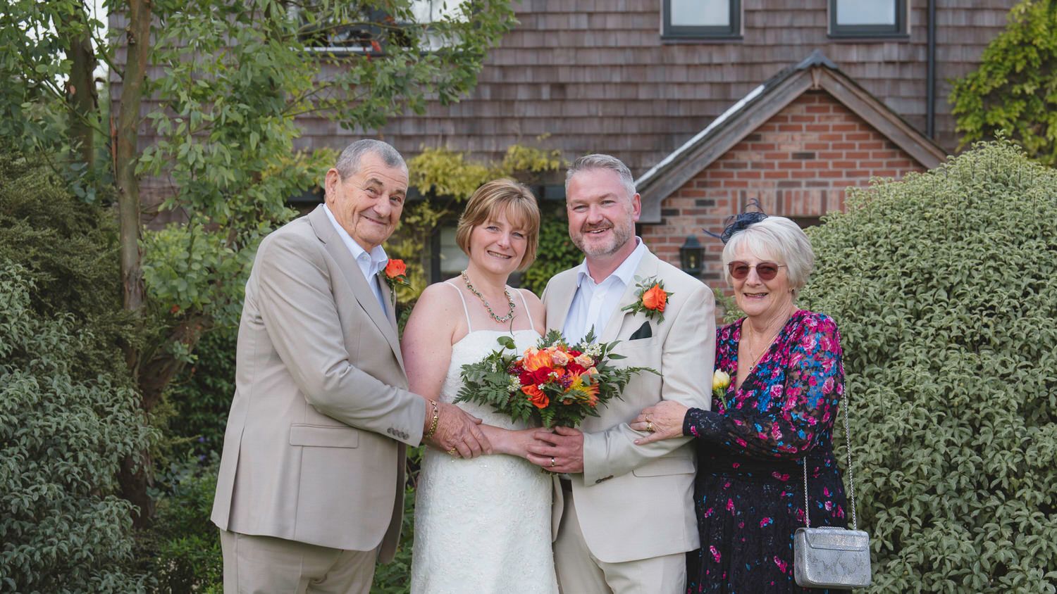 Wedding couple in white and tan suits pose with two guests outdoors, bride holding colorful bouquet.