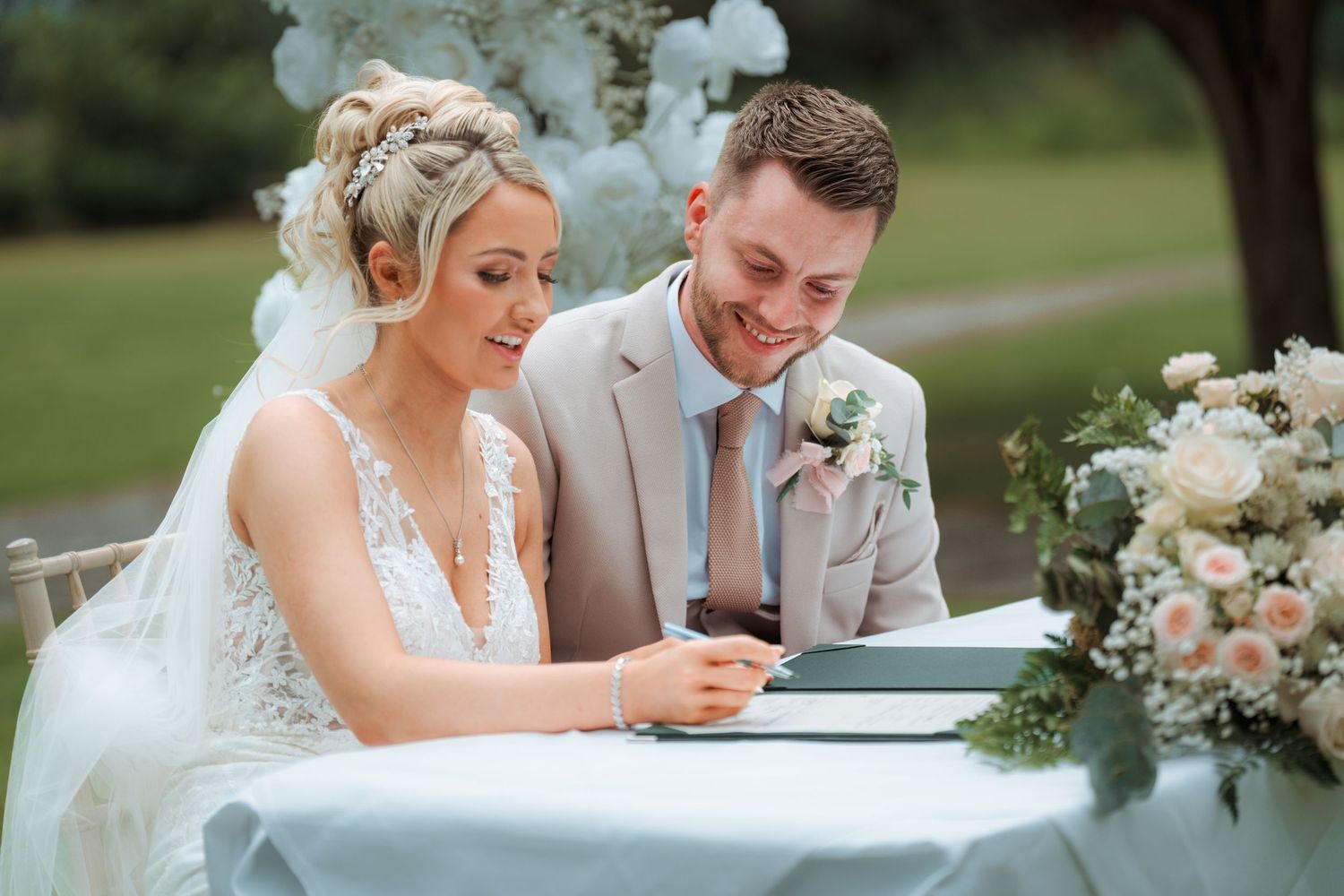 Bride and groom signing wedding register at decorated table with floral arrangements outdoors.