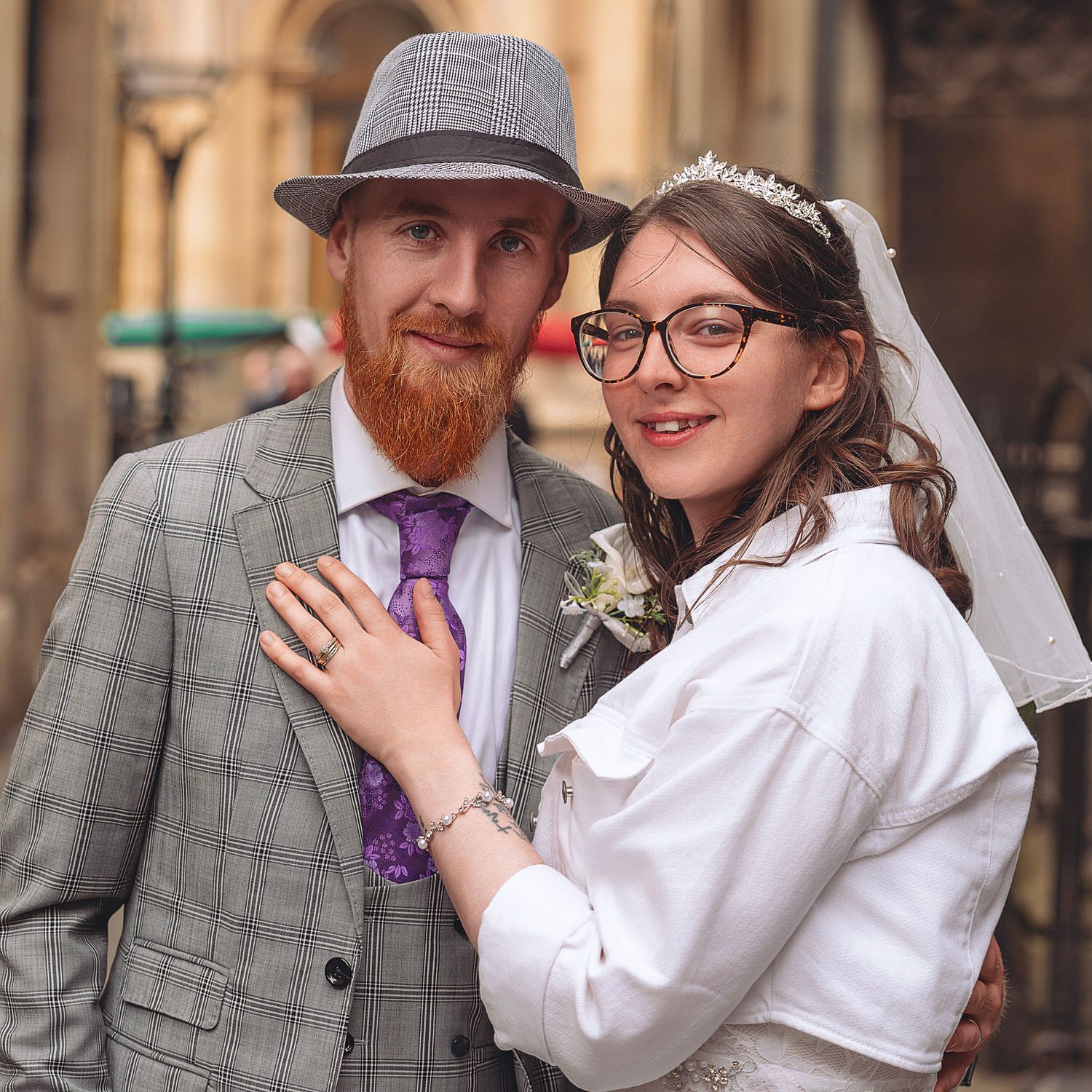 Groom in gray plaid suit and hat with red beard stands with bride in white lace dress and veil outdoors.
