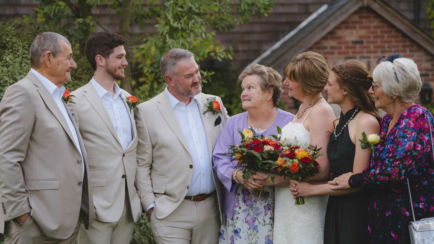Wedding group photo with bride holding colorful bouquet, surrounded by family members in formal attire outdoors.