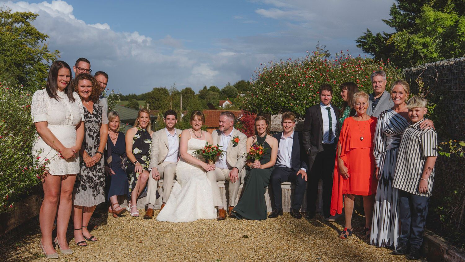 Wedding group photo with bride and groom surrounded by family and friends outdoors on a sunny day.