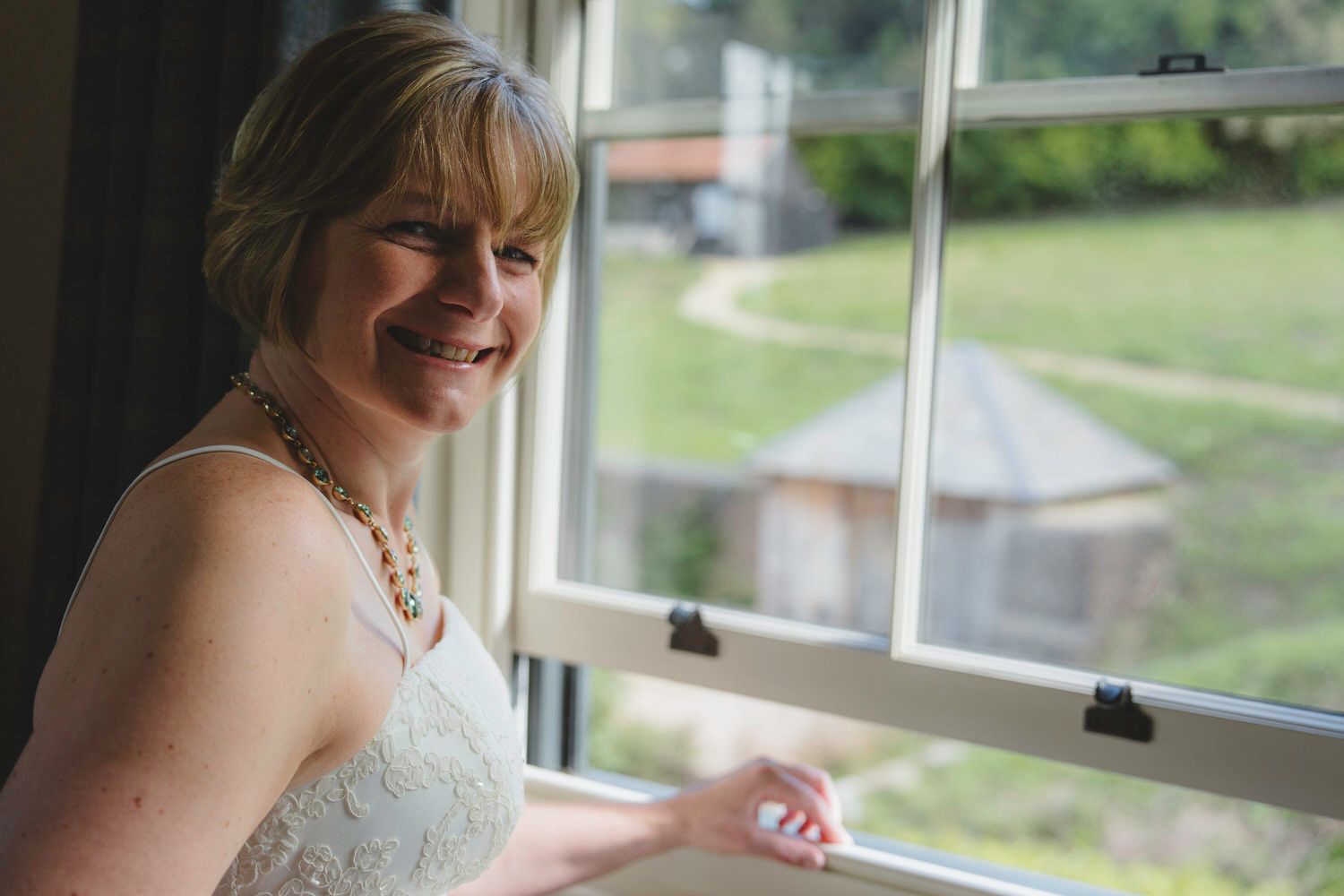 Smiling bride in lace dress standing by a window with natural light, wearing a pearl necklace.