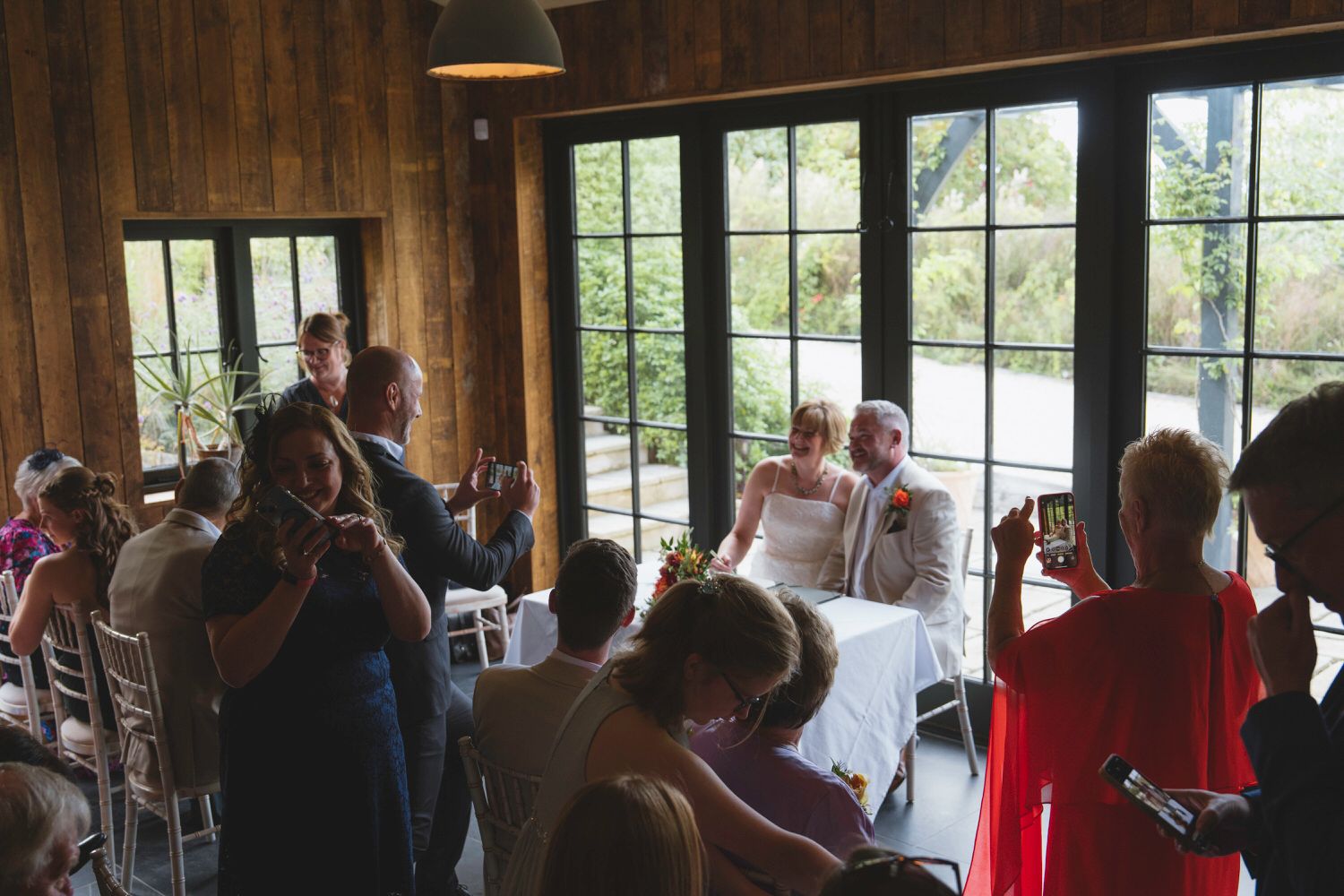 Bride and groom seated at wedding ceremony in rustic wooden venue with guests photographing the couple.