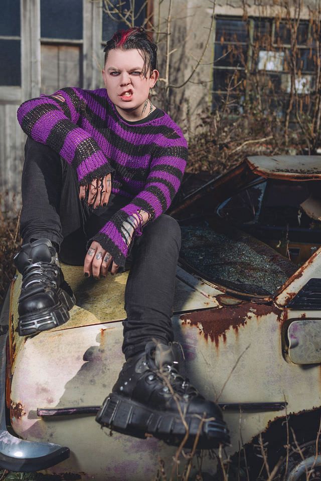 Person in torn purple striped sweater and black boots sitting on rusty abandoned car in urban decay setting.