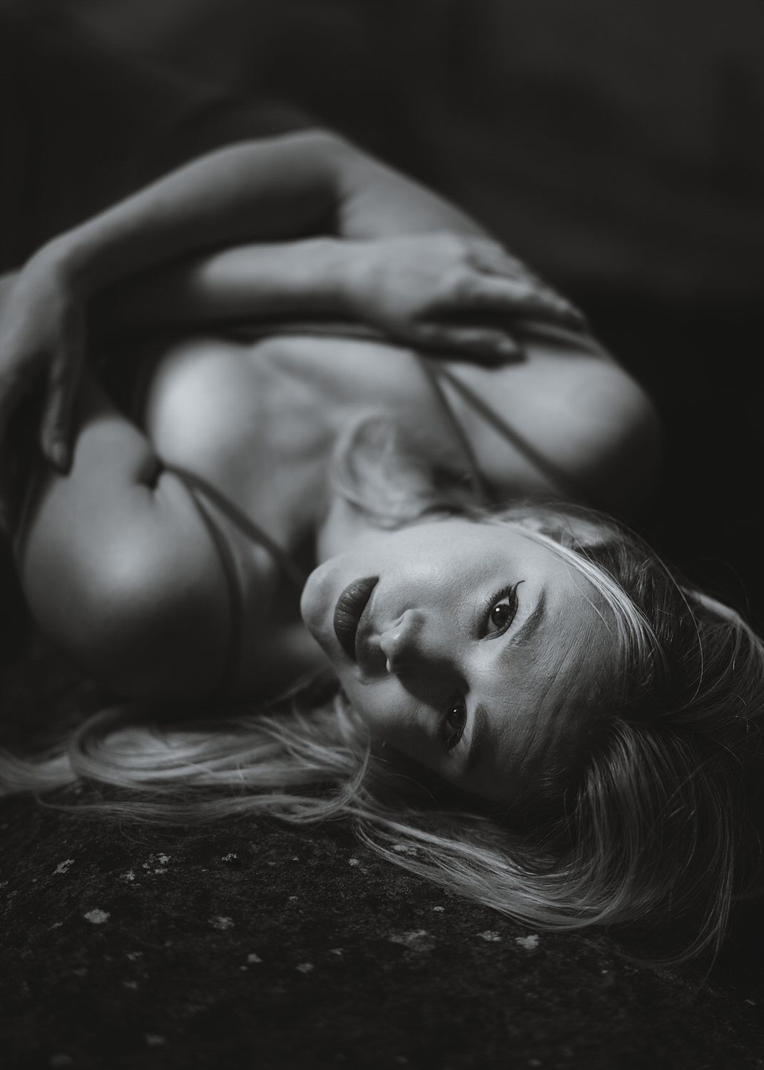 Black and white artistic boudoir photo of a blonde woman lying down with dramatic lighting and shadows.