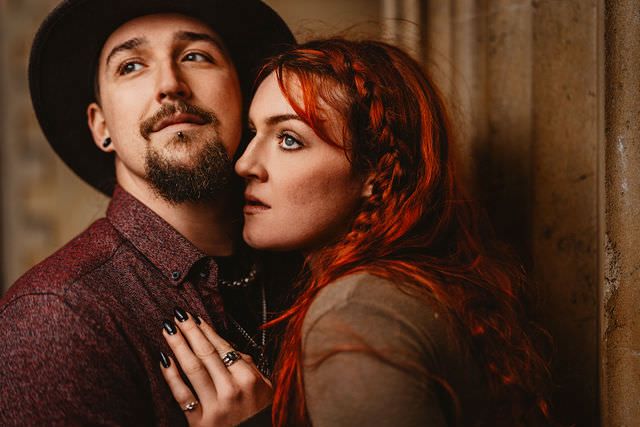 A couple poses intimately, man in black hat and woman with vibrant red braided hair leaning against stone column.