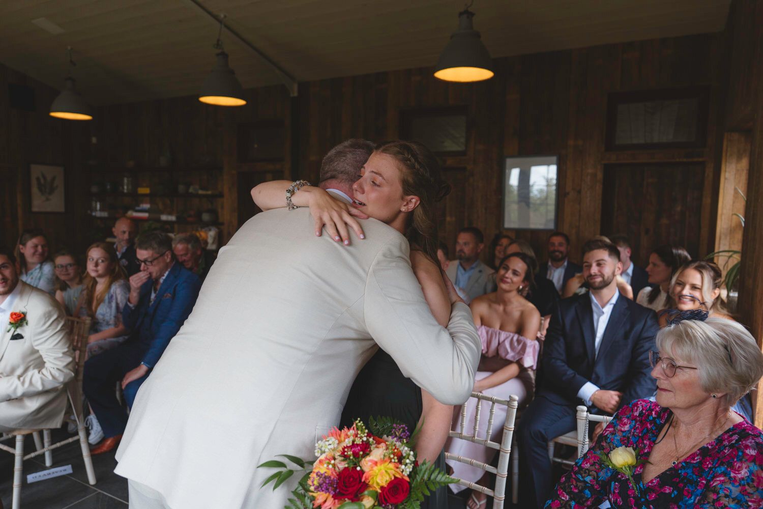 Couple embracing at wedding ceremony with guests seated in rustic venue, bride holding colorful bouquet.