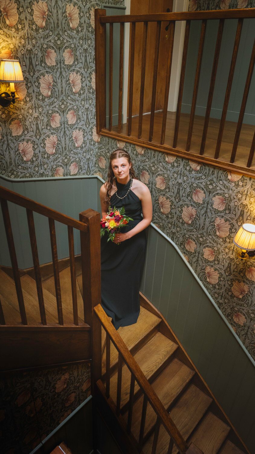 Elegant bridesmaid in black halter gown holding colorful bouquet on wooden spiral staircase with floral wallpaper.