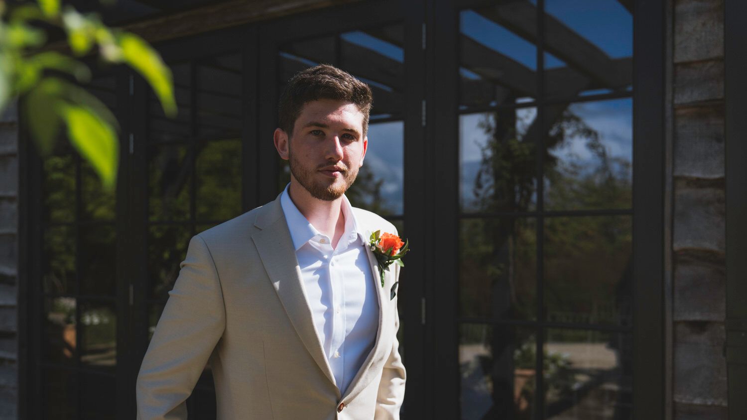 Groom in beige suit with floral boutonniere standing outdoors at wedding venue.