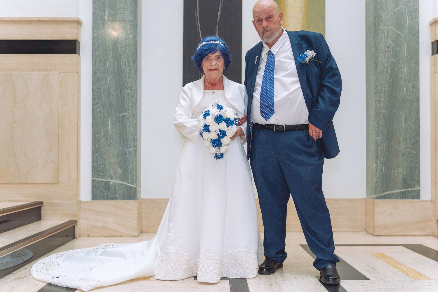 Elderly couple in wedding attire, bride in white gown with blue bouquet, groom in navy suit, posing in elegant hall.