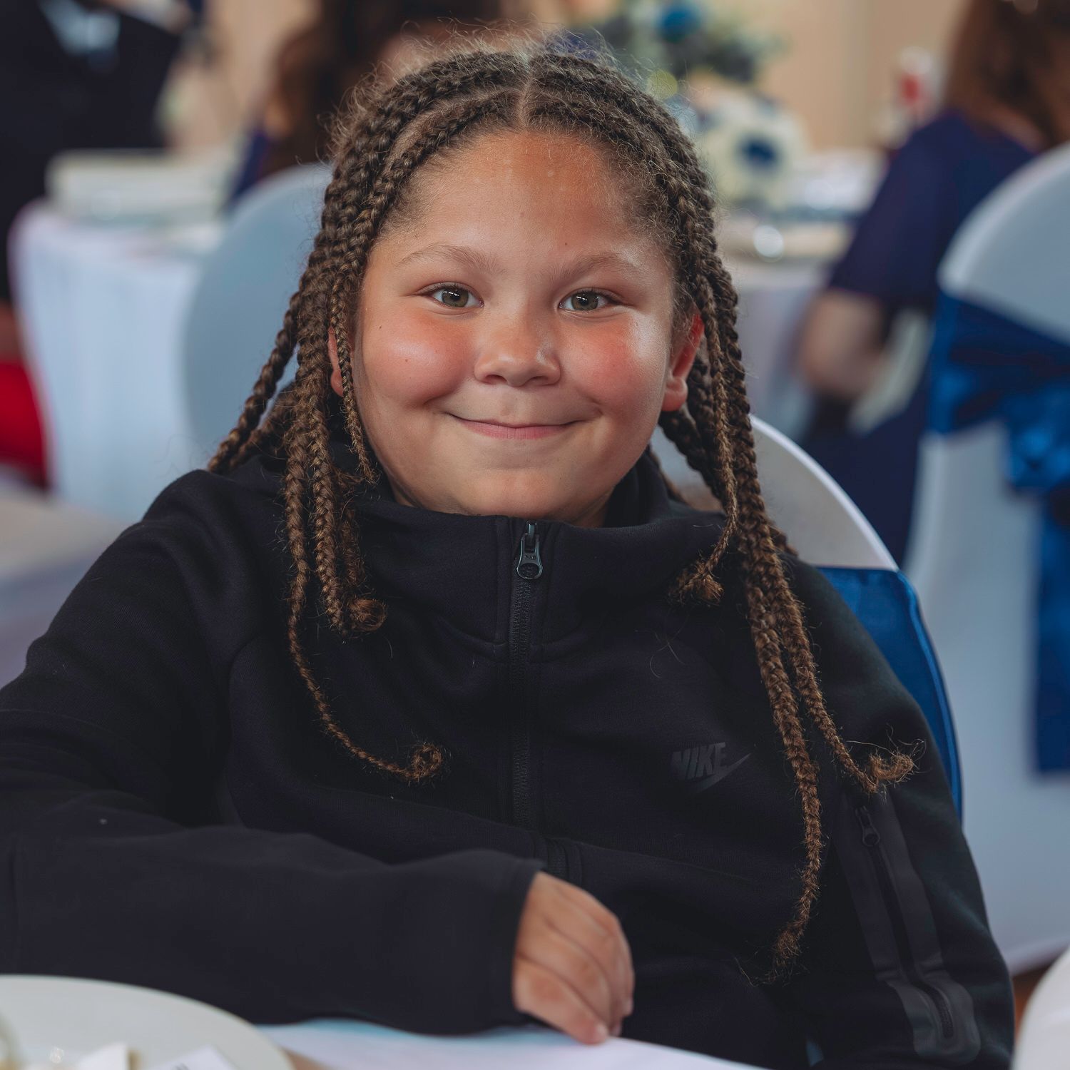 Smiling young girl with braided hair wearing a black jacket seated at a table at an indoor event.
