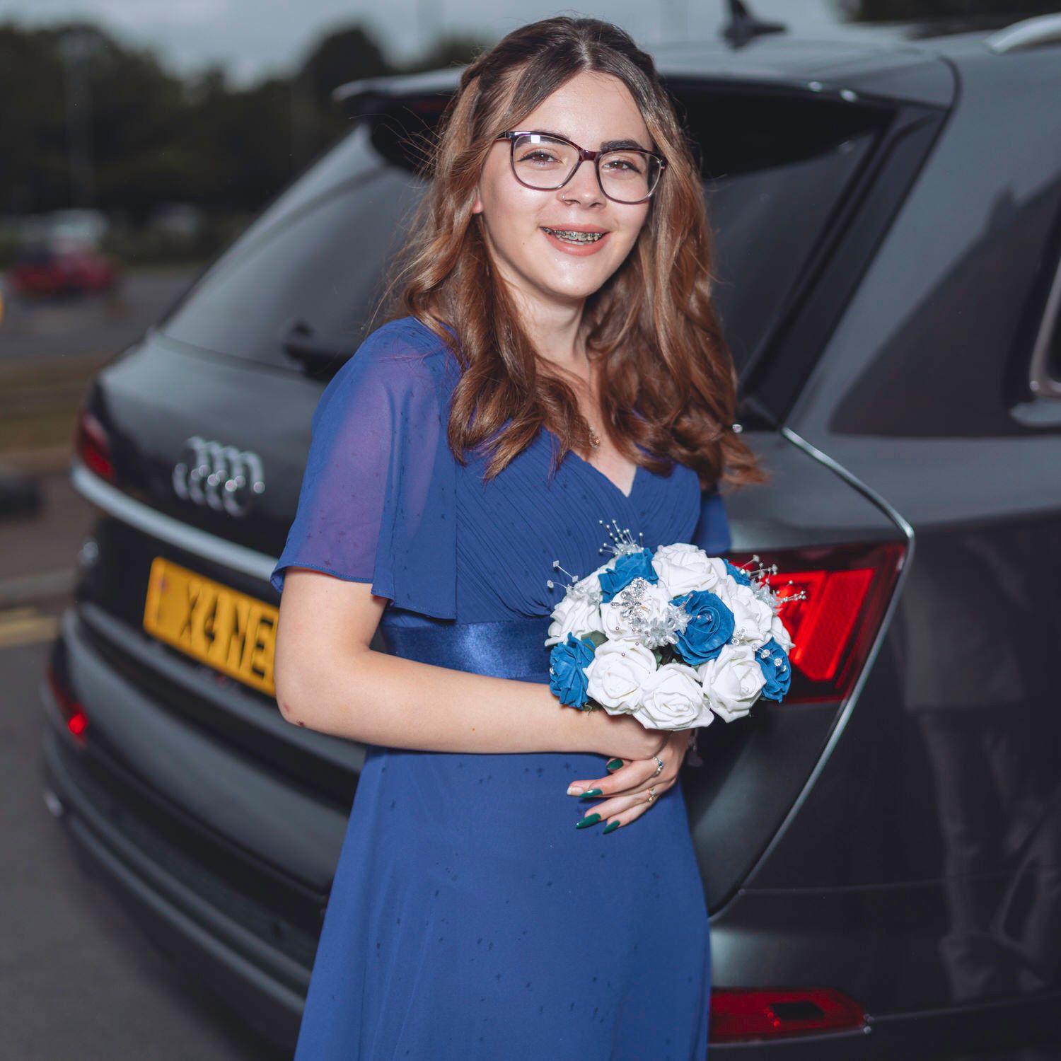 Woman in blue dress holding white and teal bouquet, standing beside a dark Audi car.