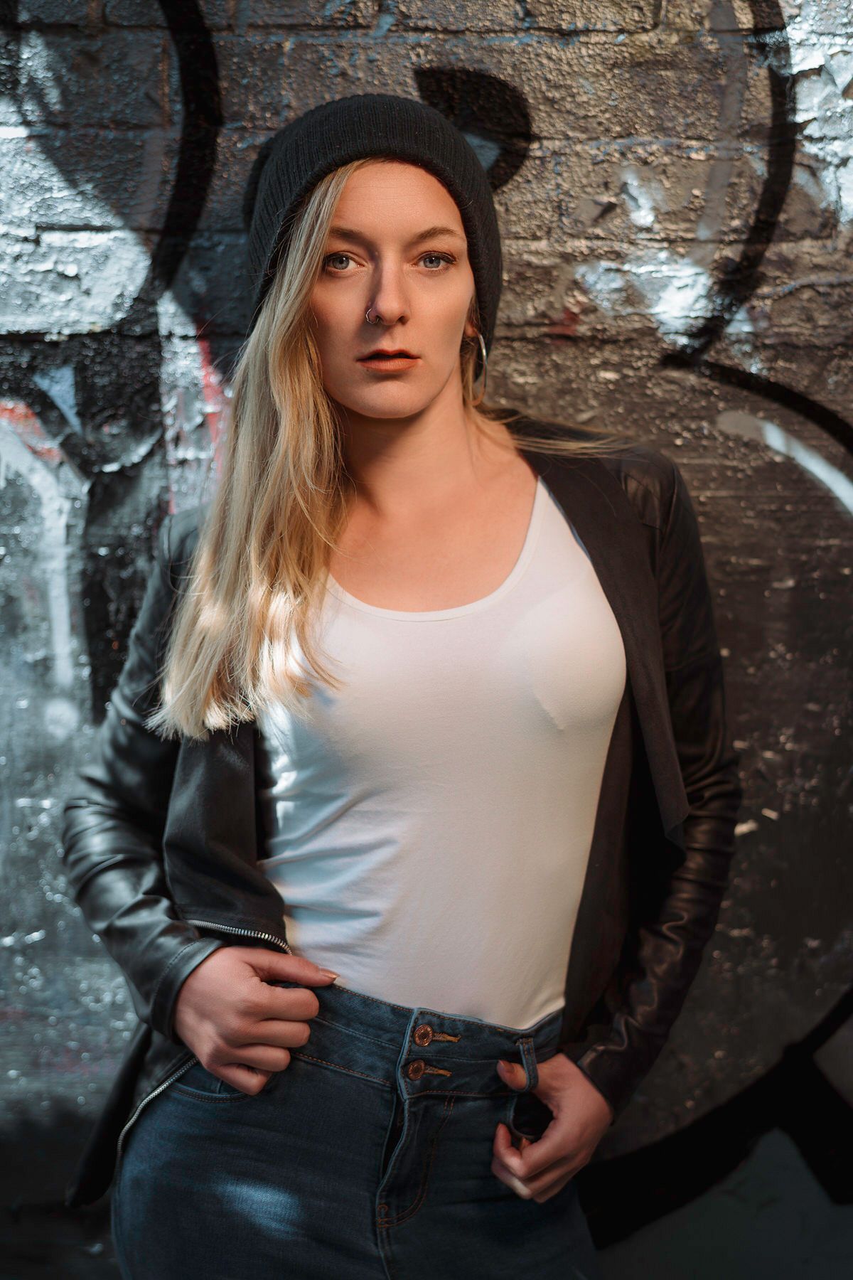 Blonde woman in white top, leather jacket and beanie hat posing against graffiti wall.