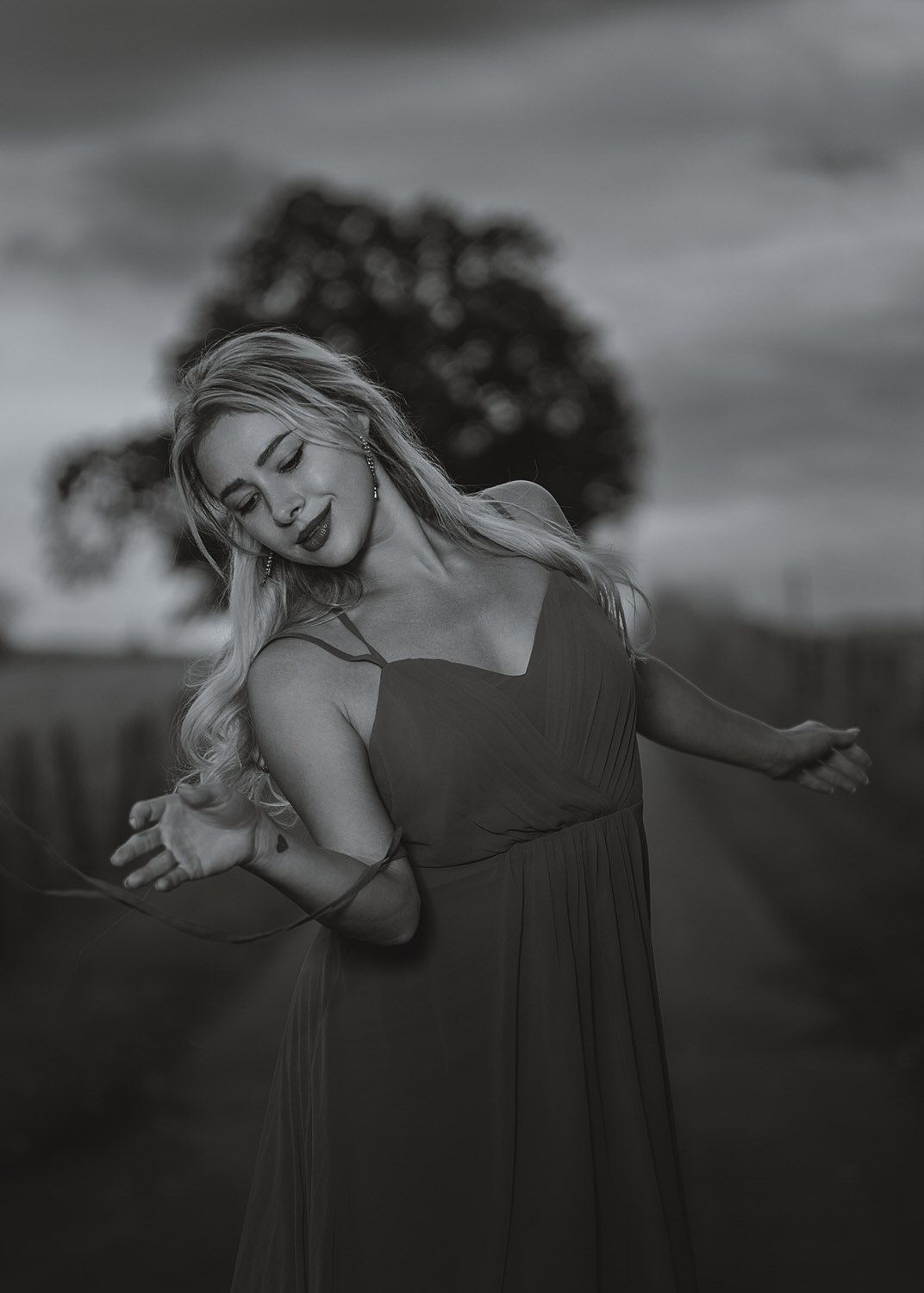 Black and white photo of a blonde woman in a flowing dress dancing gracefully outdoors near a tree at dusk.