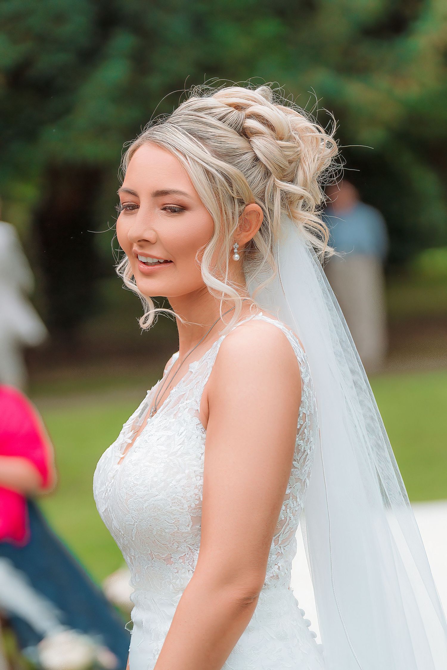 Blonde bride in white lace gown and veil with elegant updo hairstyle smiling outdoors at wedding ceremony.