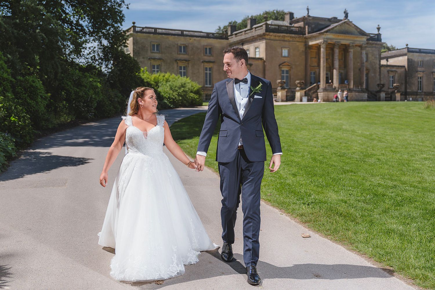 Happy newlyweds walk hand in hand along path with grand historic manor house behind them.
