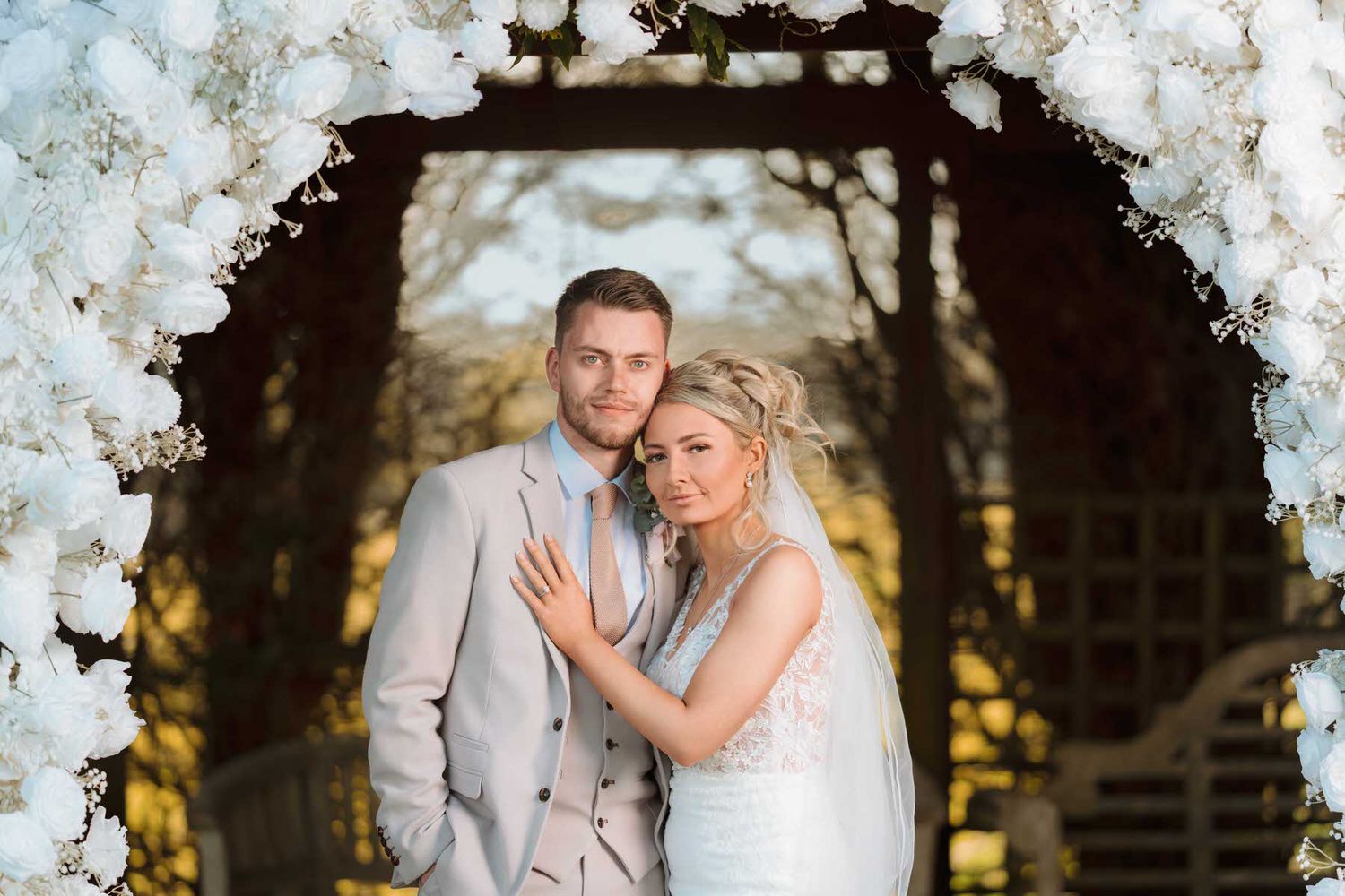 Bride and groom posing under white floral arch in golden autumn garden setting.