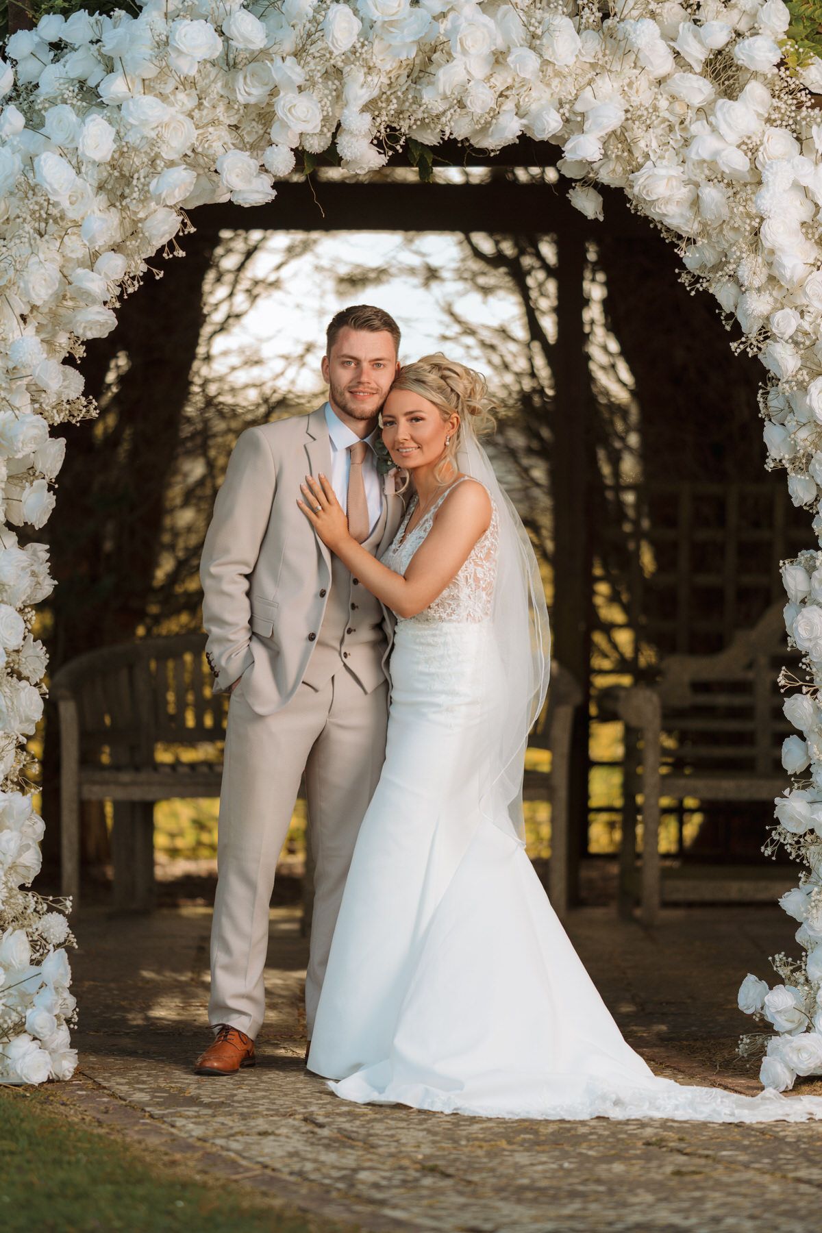 Bride and groom posing under a white floral arch in a garden, wearing a wedding dress and gray suit.