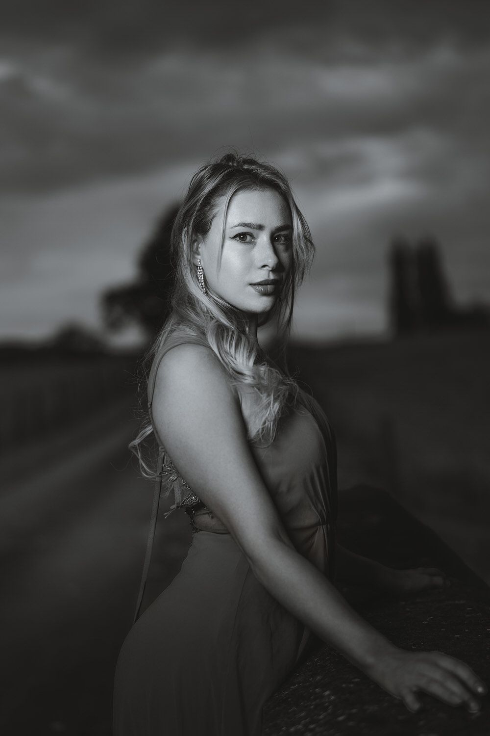 Black and white portrait of a blonde woman in a dress posing outdoors on a rural road under dramatic cloudy skies.