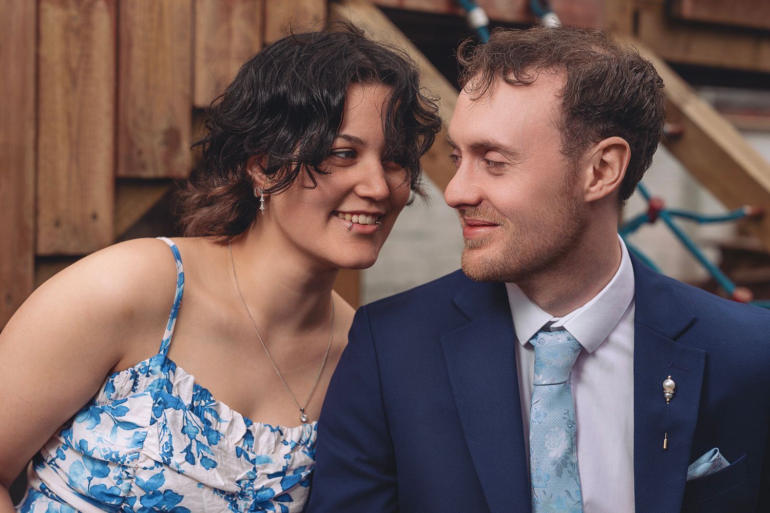 A smiling couple in formal attire share a romantic moment, woman in floral dress and man in navy suit with teal tie.