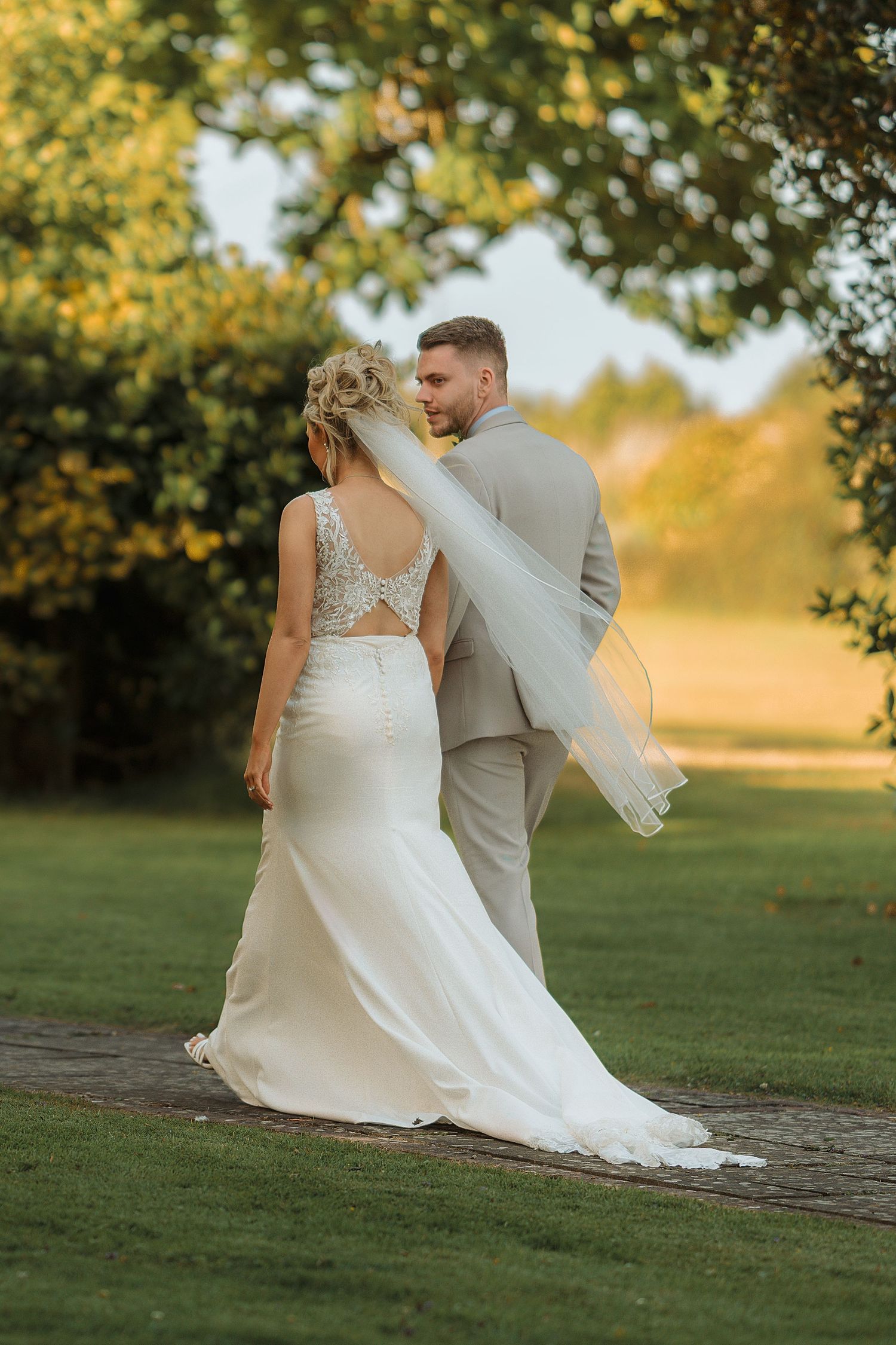 Bride in white gown and veil walks with groom in grey suit through lush garden at golden hour.