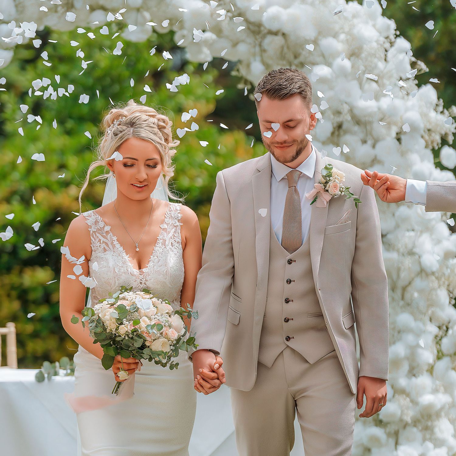 A bride and groom walk hand in hand under a white floral arch with confetti falling at their outdoor wedding ceremony.