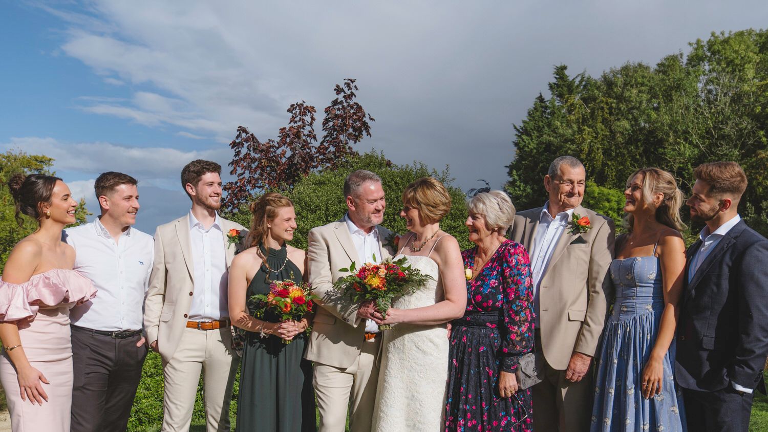 Wedding party group photo outdoors, bride and groom with family smiling, lush greenery and cloudy sky backdrop.