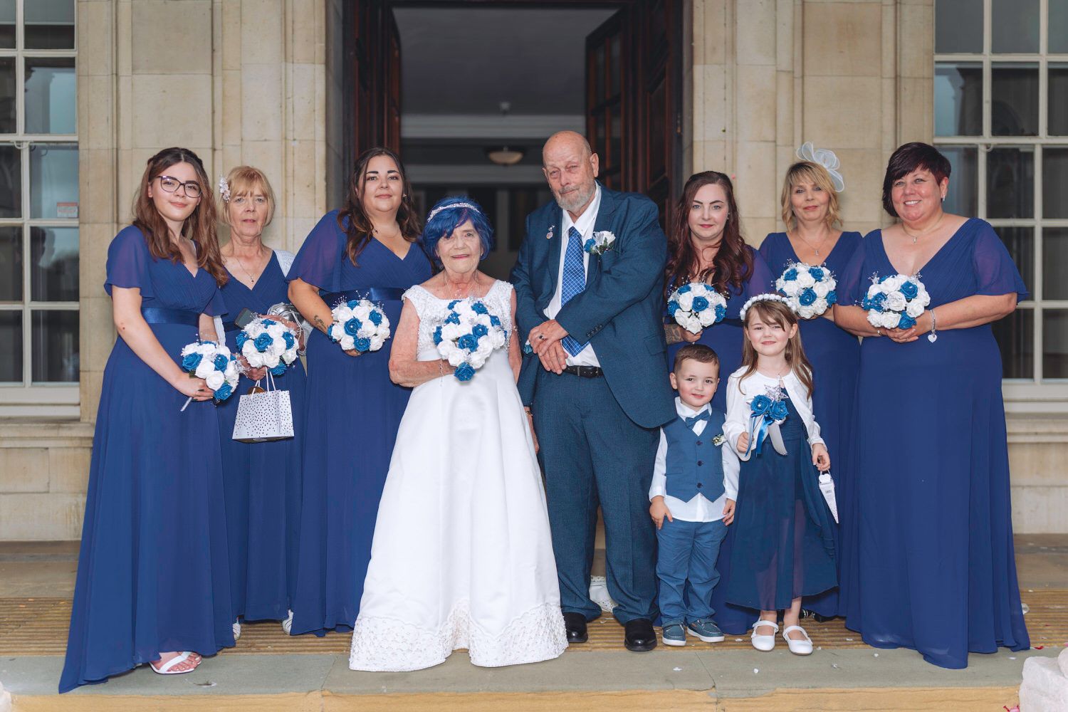 Wedding party posing in navy blue, bride in white gown, groom in suit, bridesmaids holding blue and white bouquets.