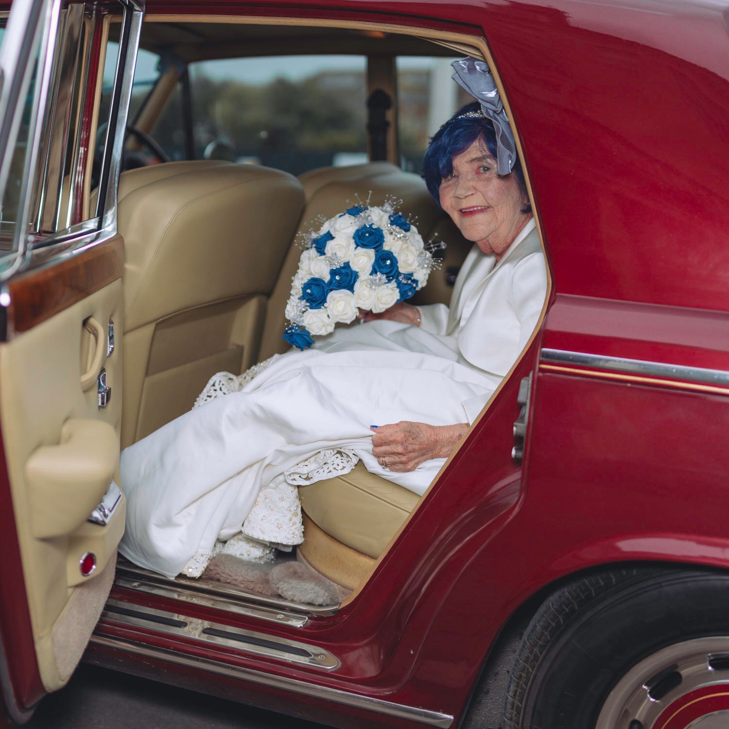 Elderly bride in white gown with blue bouquet sitting in classic red car on wedding day.