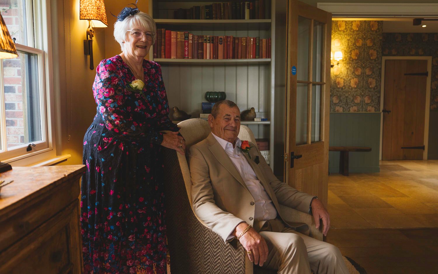 Elderly couple posing in cozy living room, woman standing in floral dress beside seated man in tan suit.
