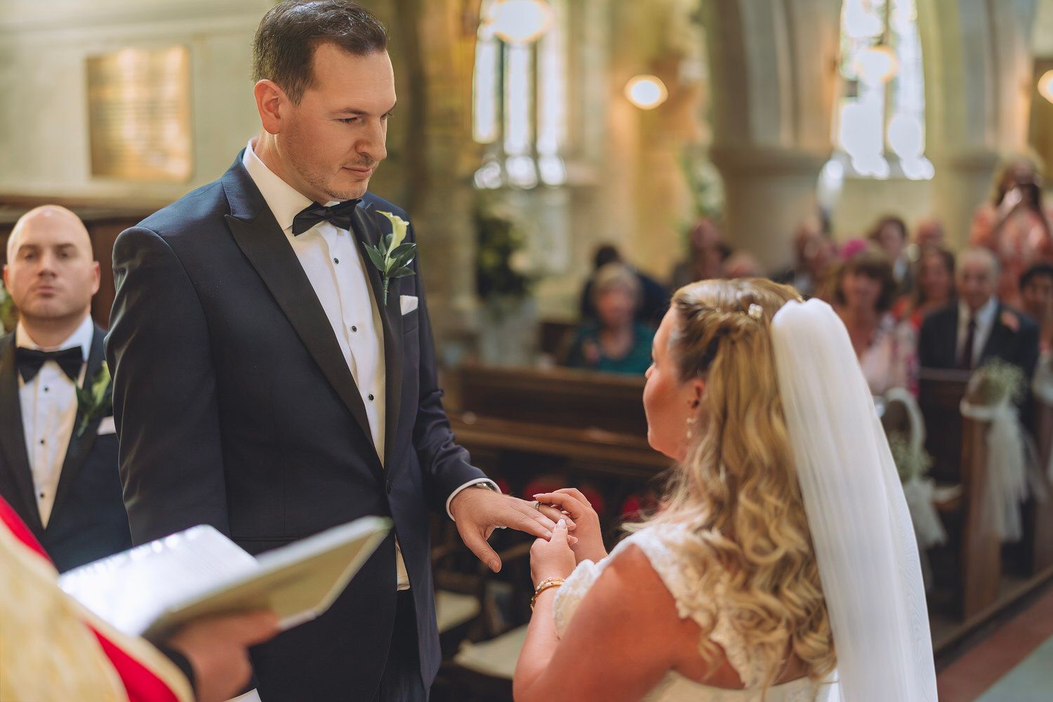 Groom placing wedding ring on bride's finger during church ceremony, with guests seated in pews in the background.