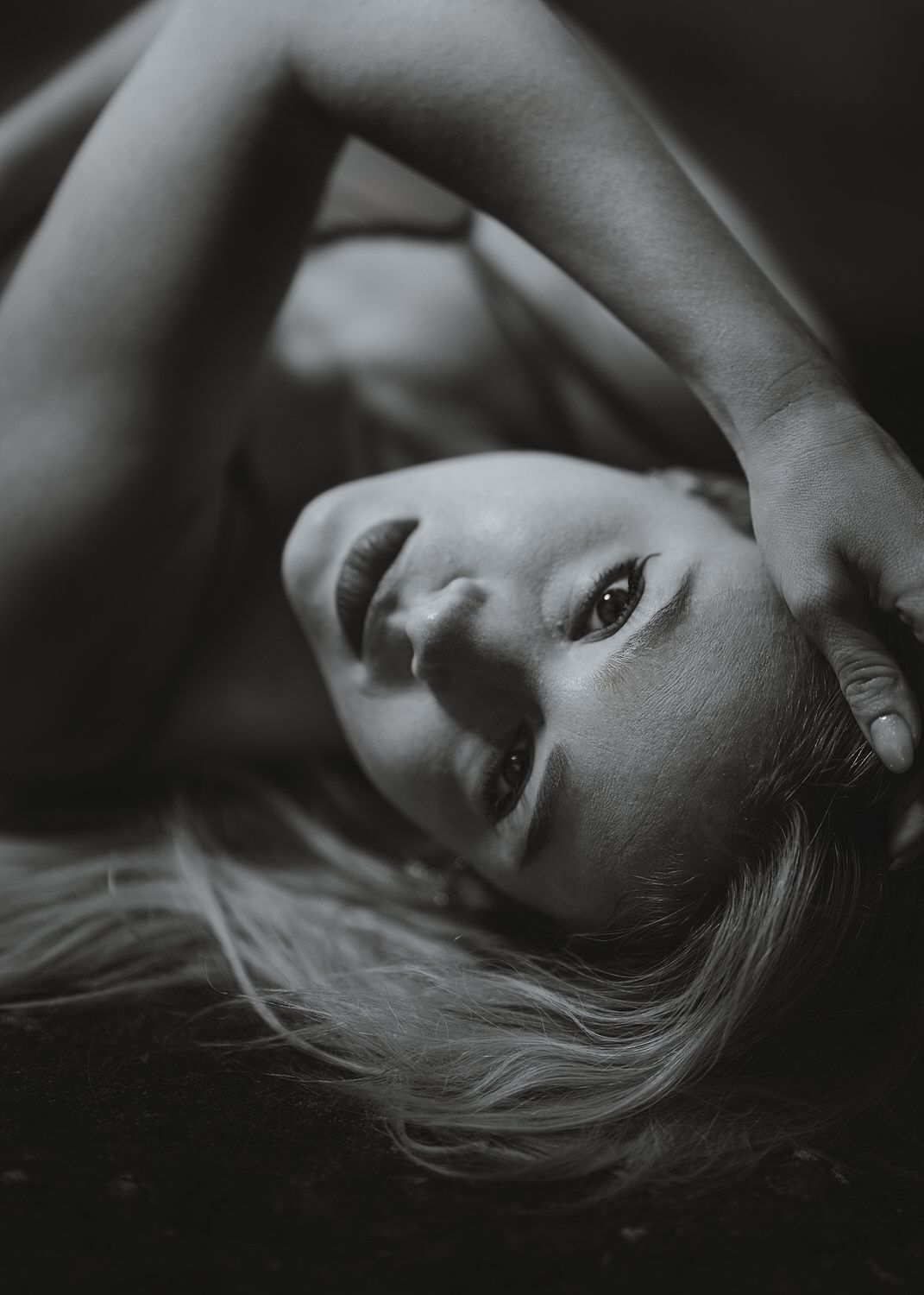 Black and white portrait of a young woman lying back with arms raised, gazing upward with long blonde hair spread out.