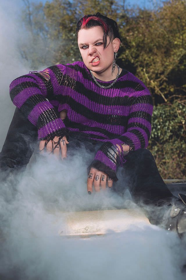 Person in purple striped sweater with tattoos poses dramatically amid smoke outdoors.