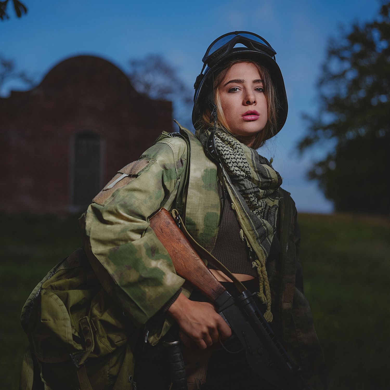 Young woman in camouflage gear holding rifle posing near old brick building at sunset.
