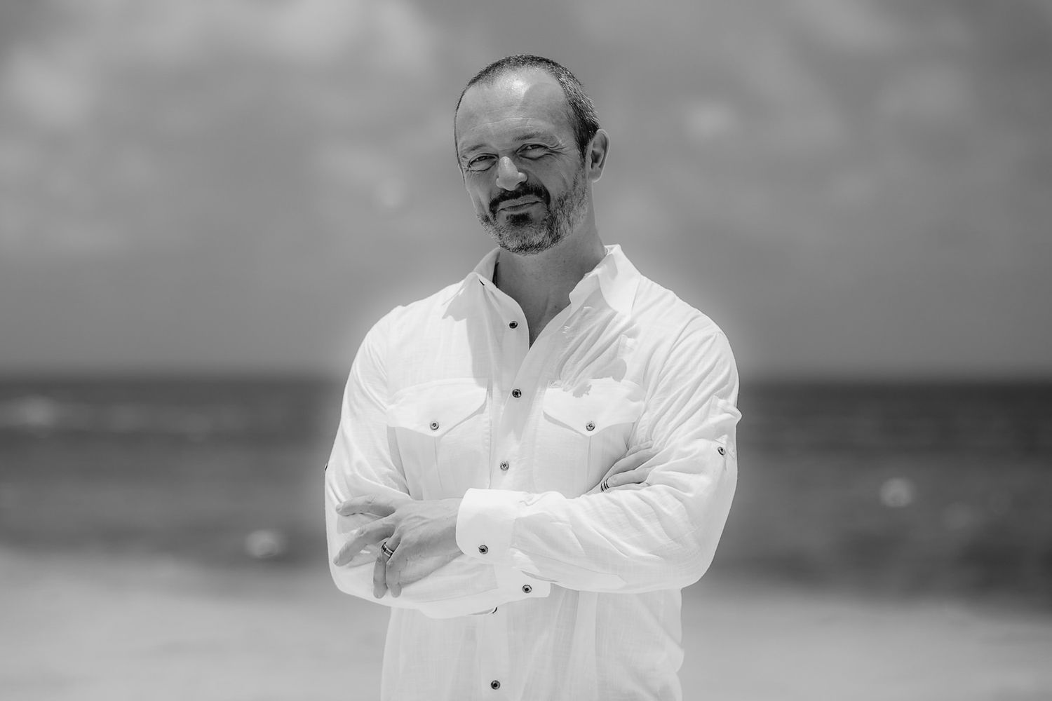 Black and white portrait of confident man in white linen shirt standing on beach with arms crossed.