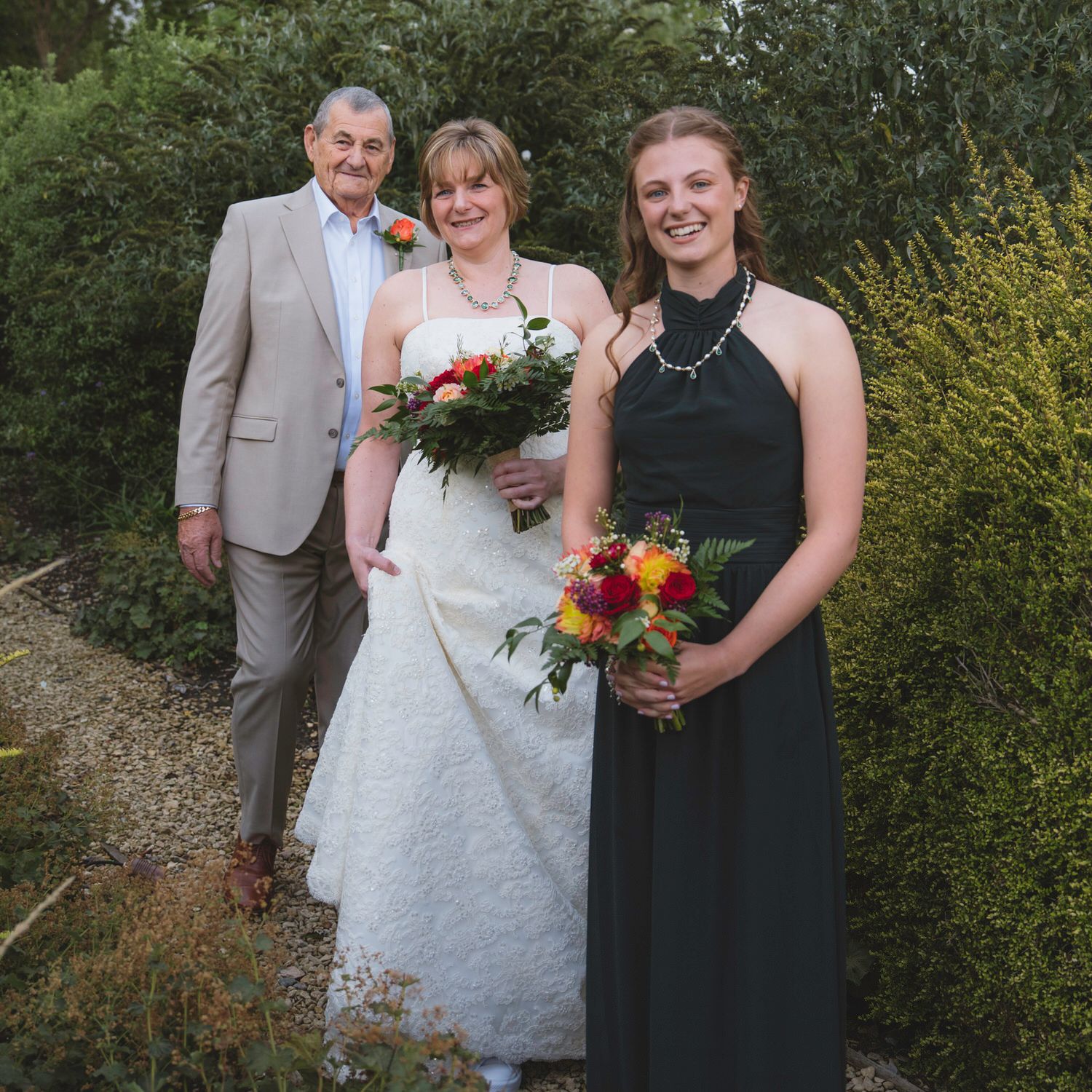 Bride in white gown with bouquet poses with bridesmaid in black dress and older gentleman at outdoor wedding.
