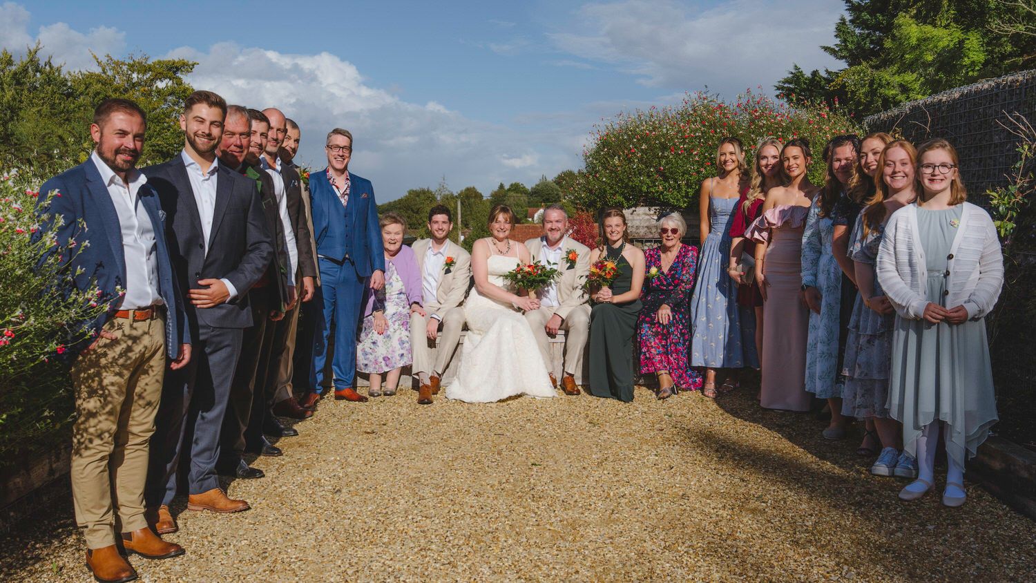 Wedding group photo with bride and groom surrounded by guests in a garden setting on a sunny day.