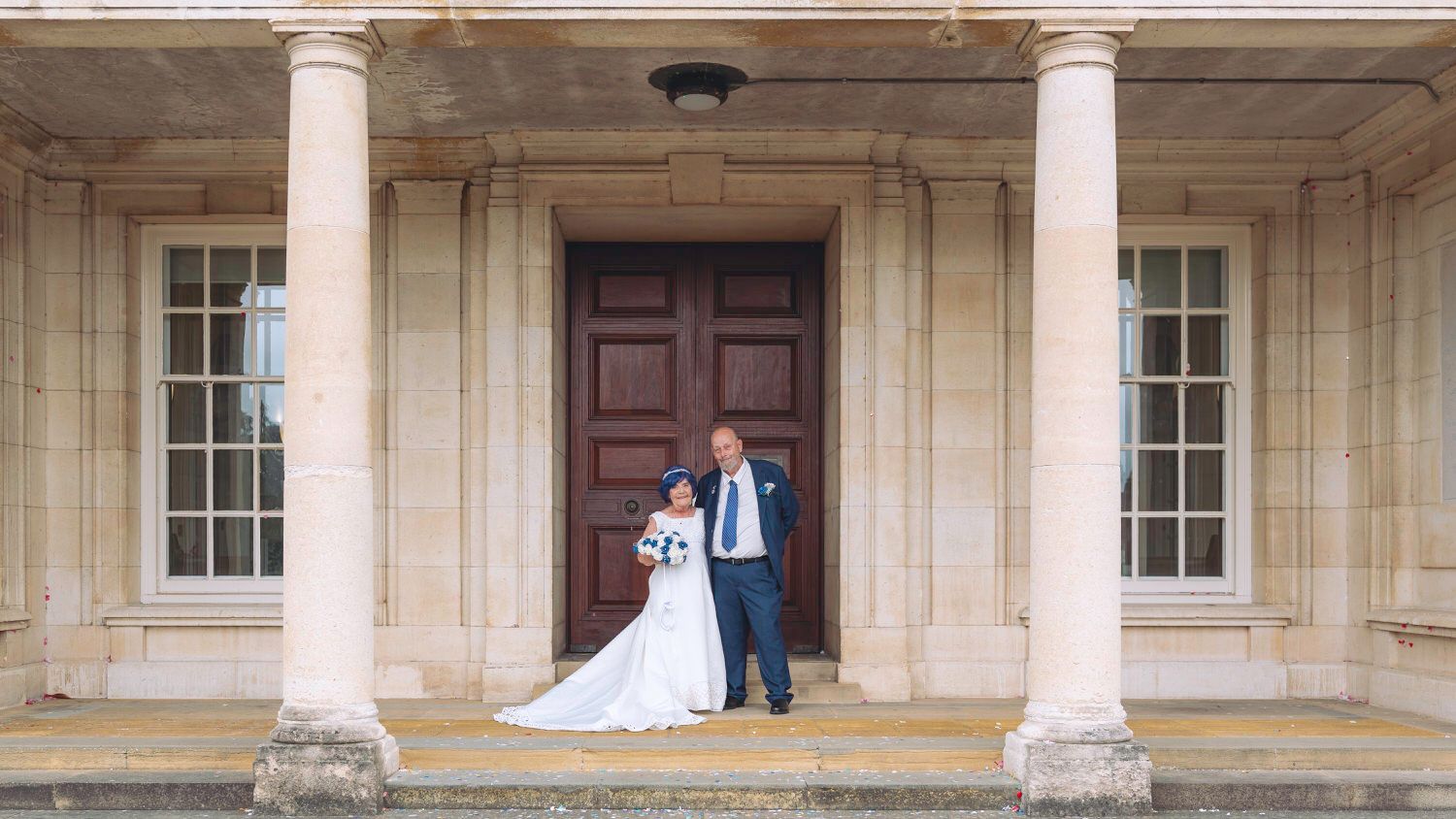 Bride and groom standing together at elegant stone building entrance with tall columns on their wedding day.