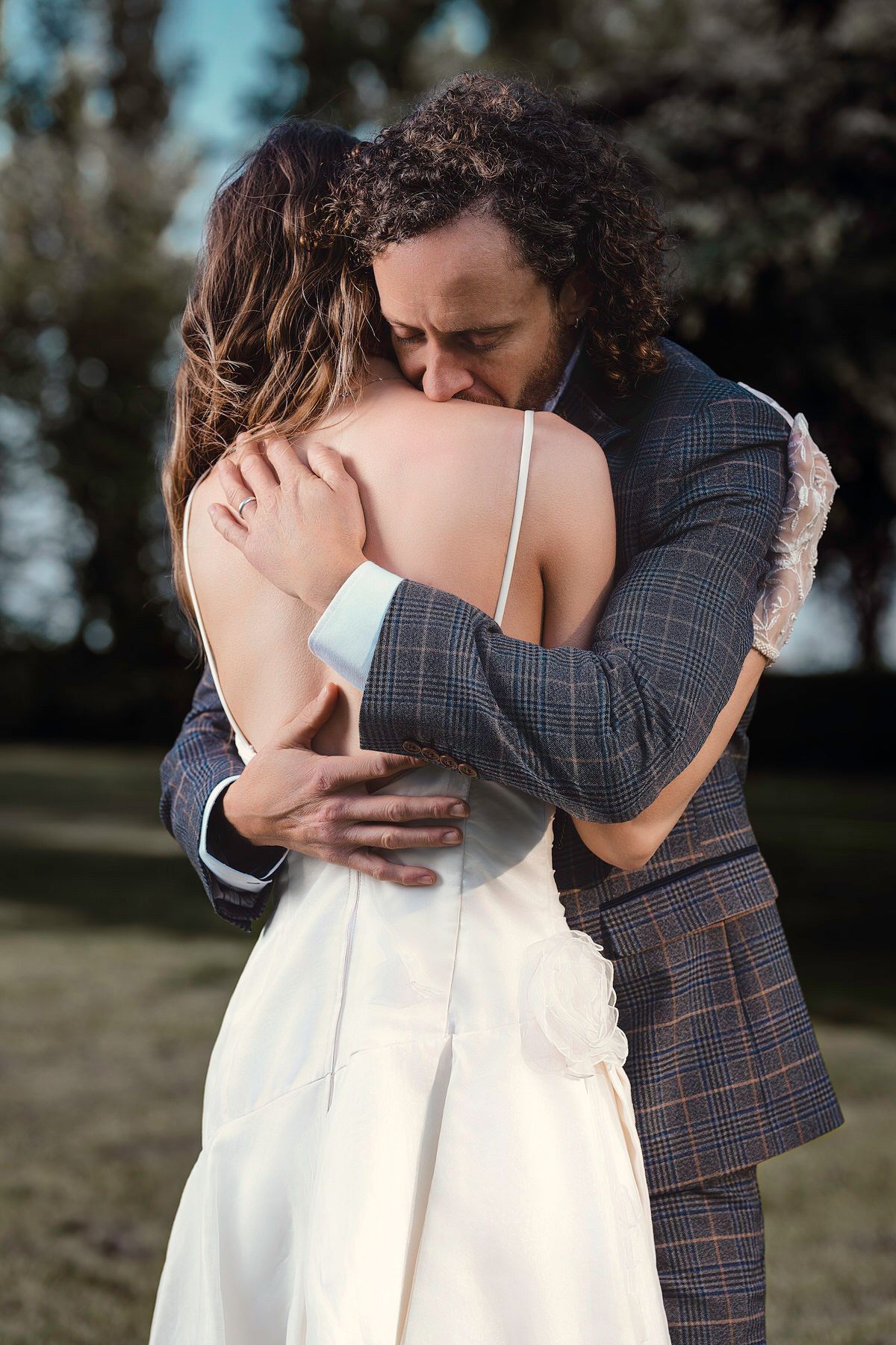 Groom in plaid suit embracing bride in white slip dress in an emotional outdoor wedding moment.