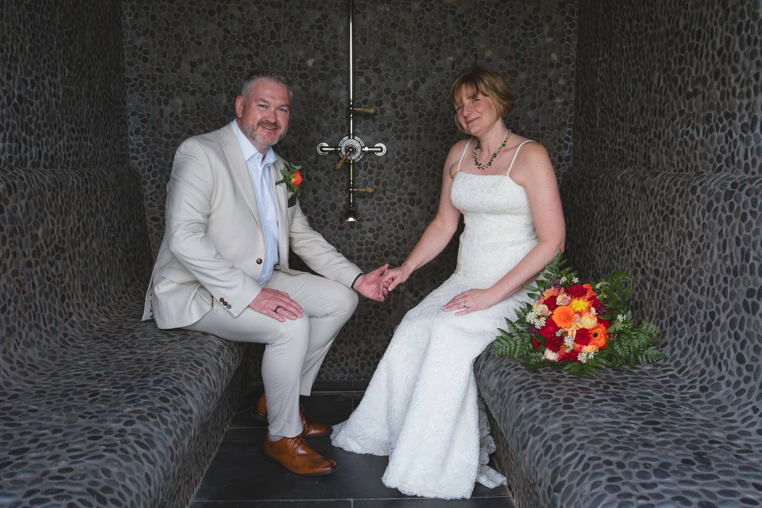 Bride and groom holding hands while seated in an elegant stone interior, bride holding a vibrant bouquet.