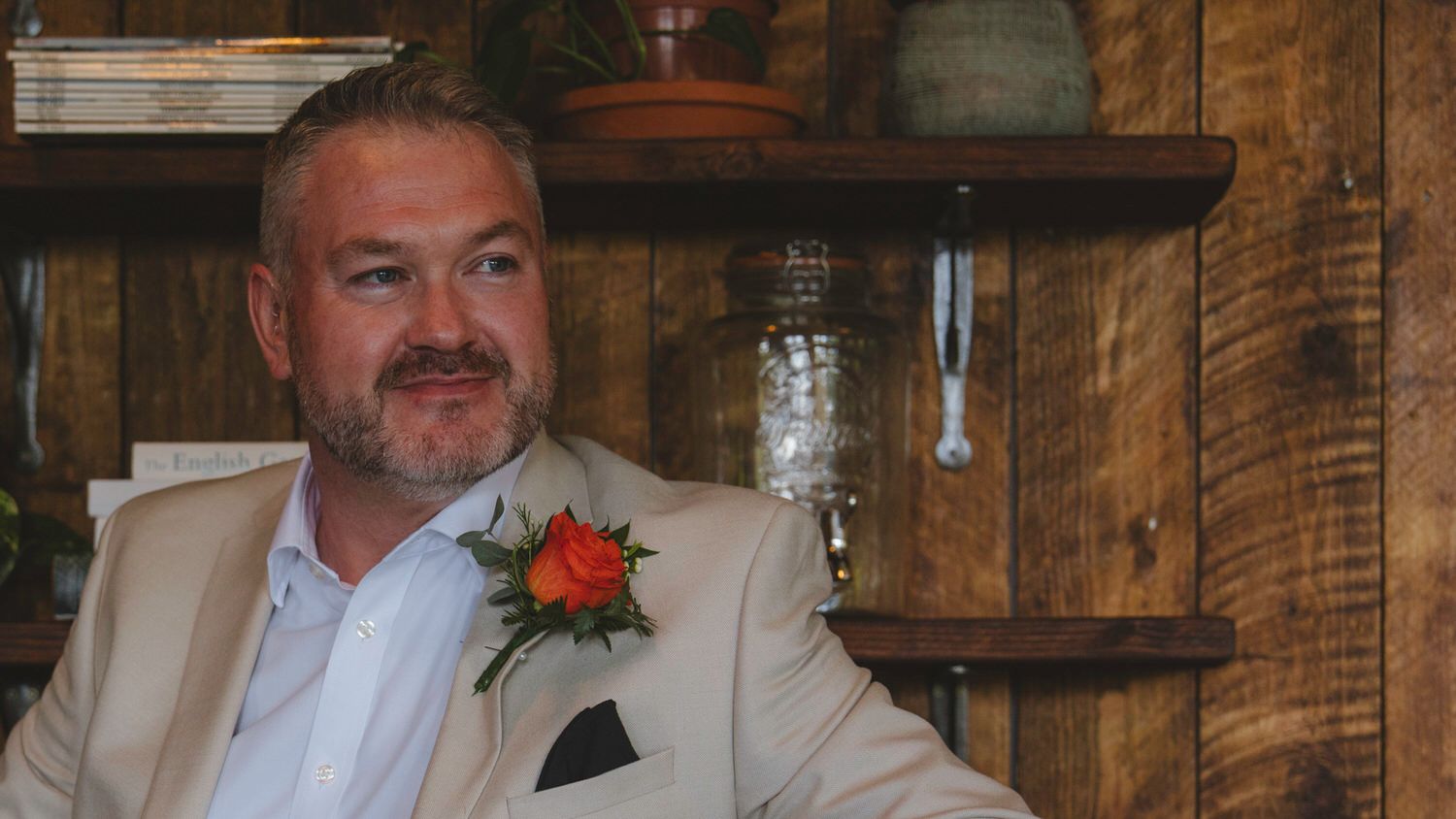 Man in beige suit with orange boutonniere smiling indoors near rustic wooden shelves.