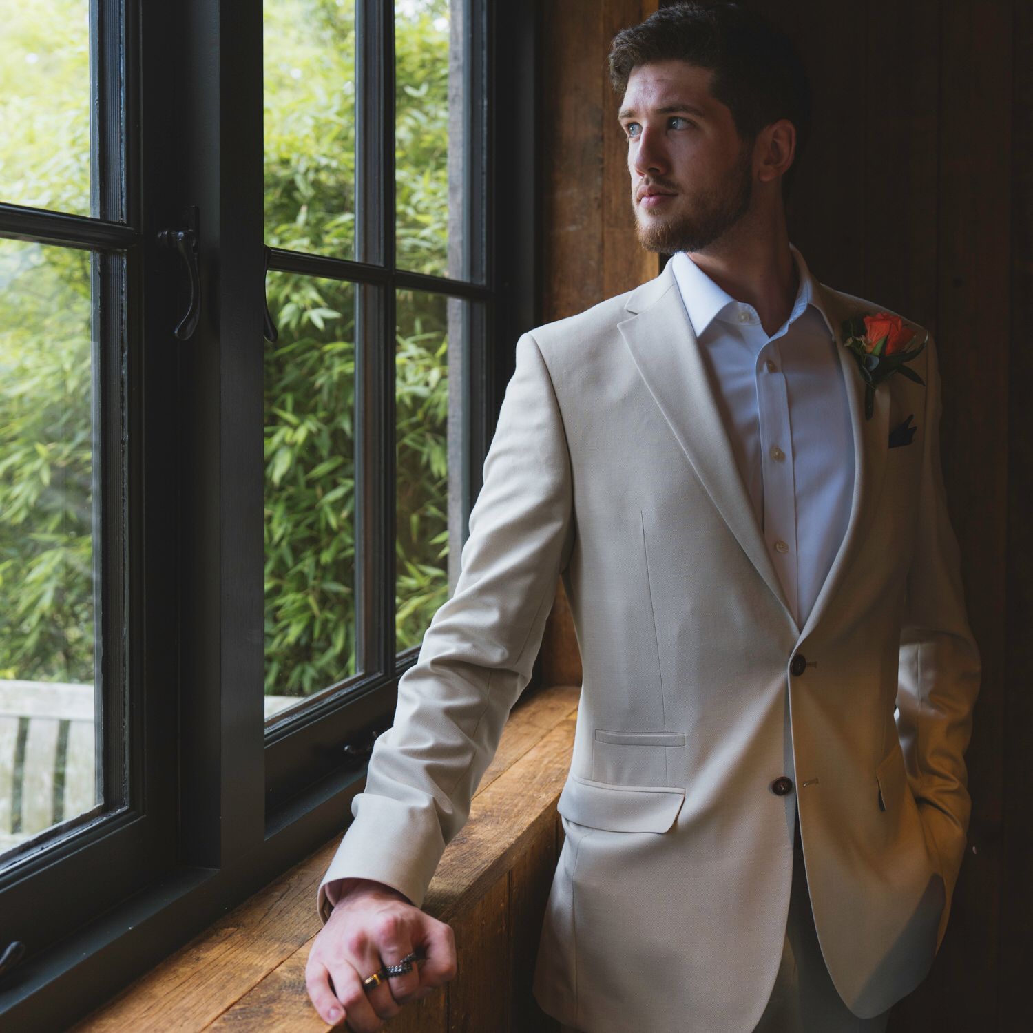 Elegant groom in a beige suit with boutonniere stands by a window, gazing outside at lush greenery.