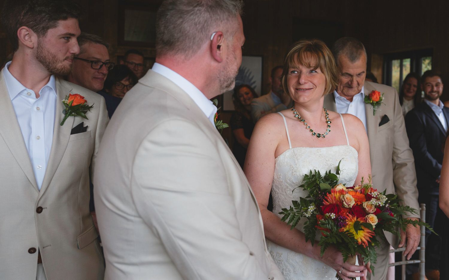 Bride smiling at groom during outdoor wedding ceremony, holding colorful bouquet of red and orange flowers.