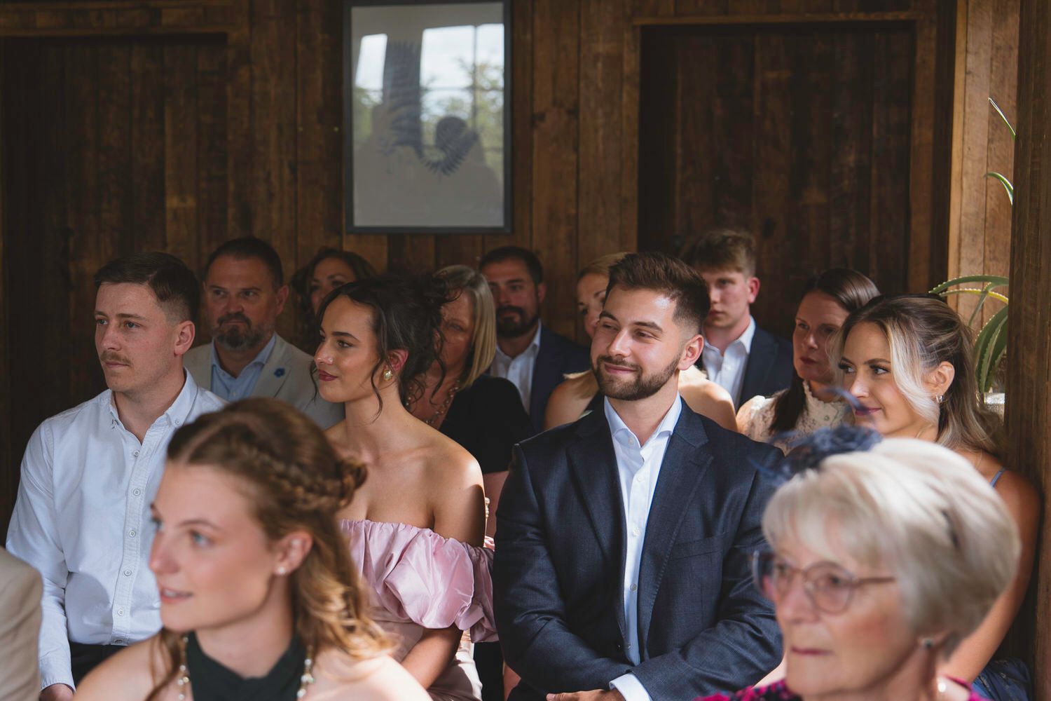 Guests seated indoors at a wedding ceremony, smiling and watching attentively in a rustic wooden venue.