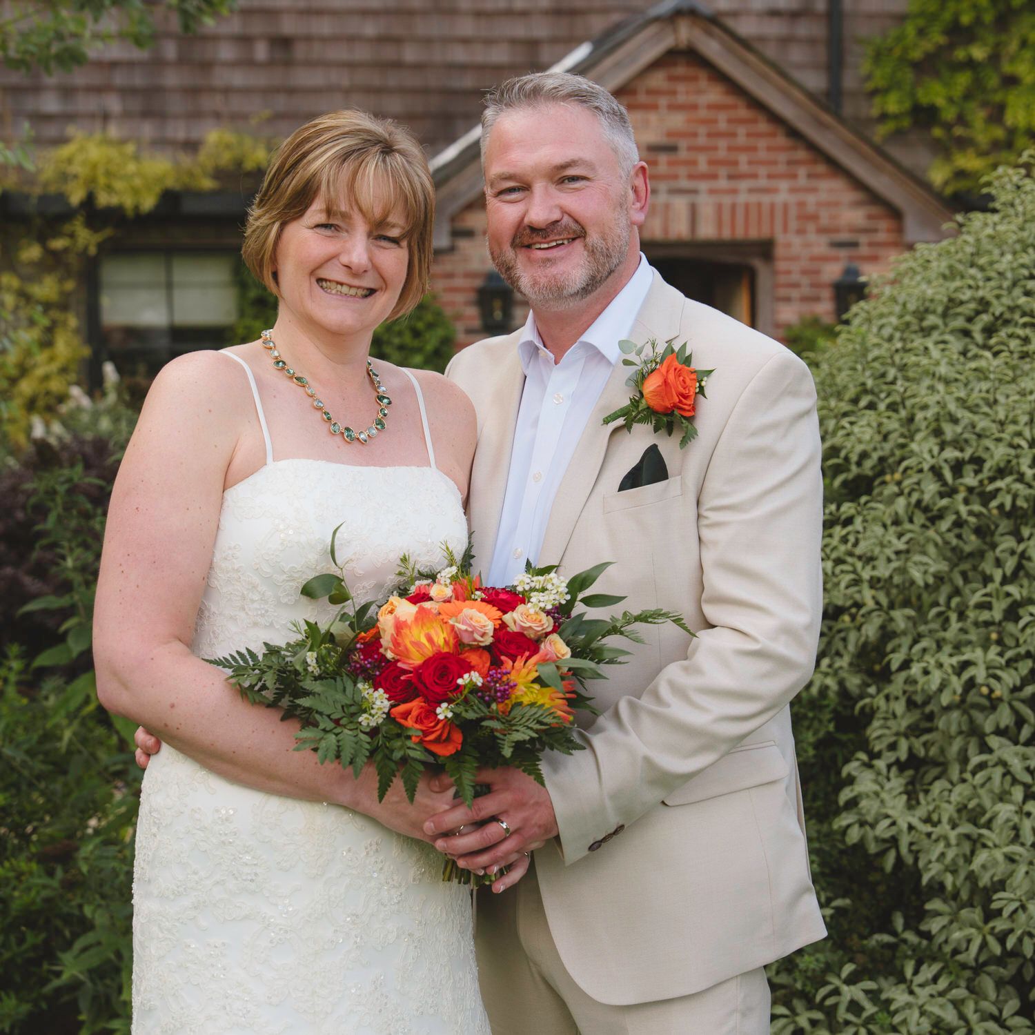 Bride in white dress holding colorful bouquet stands with groom in beige suit outside brick building.