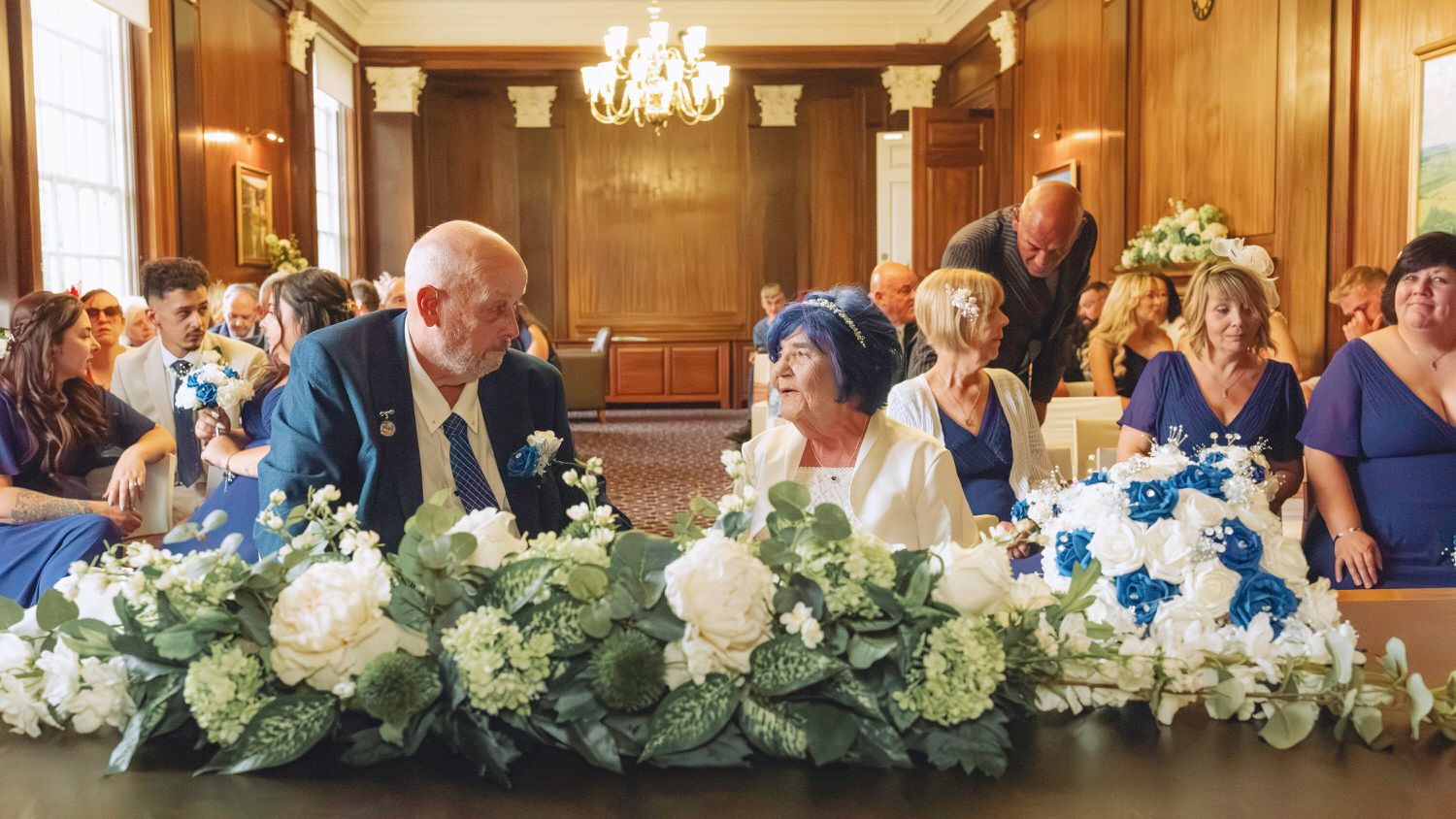 Elderly couple exchanging vows at wedding ceremony with blue and white floral arrangements in elegant venue.