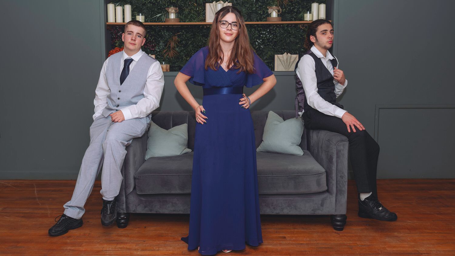 Woman in blue gown poses between two men in suits on a gray sofa in an elegant room.