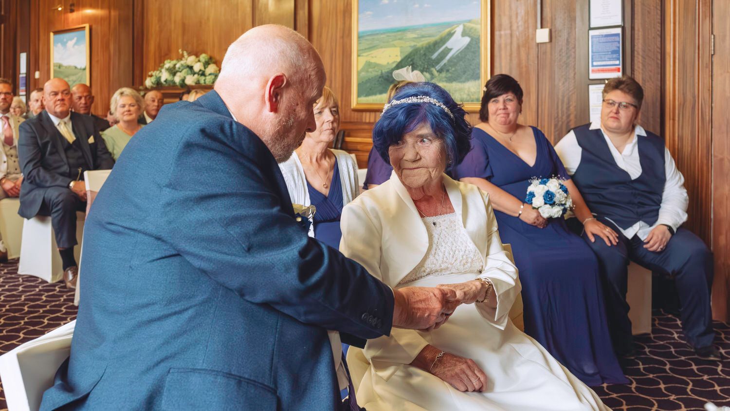 Elderly couple exchanging vows at an indoor wedding ceremony with guests and bridesmaids in the background.