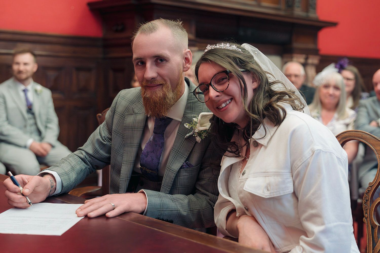 Happy couple signing marriage register at civil wedding ceremony, bride in white with floral headpiece, groom with red beard.